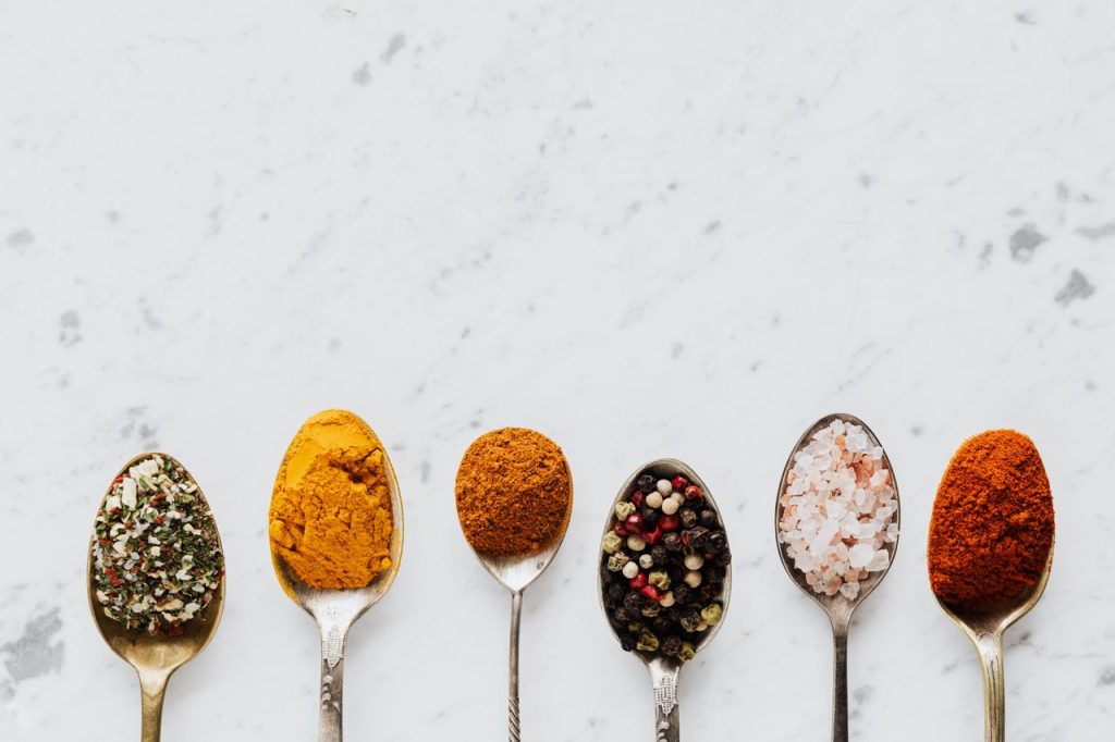 Top view of assorted aromatic condiments arranged together in metal spoons on marble surface used for delicious gourmet meal cooking